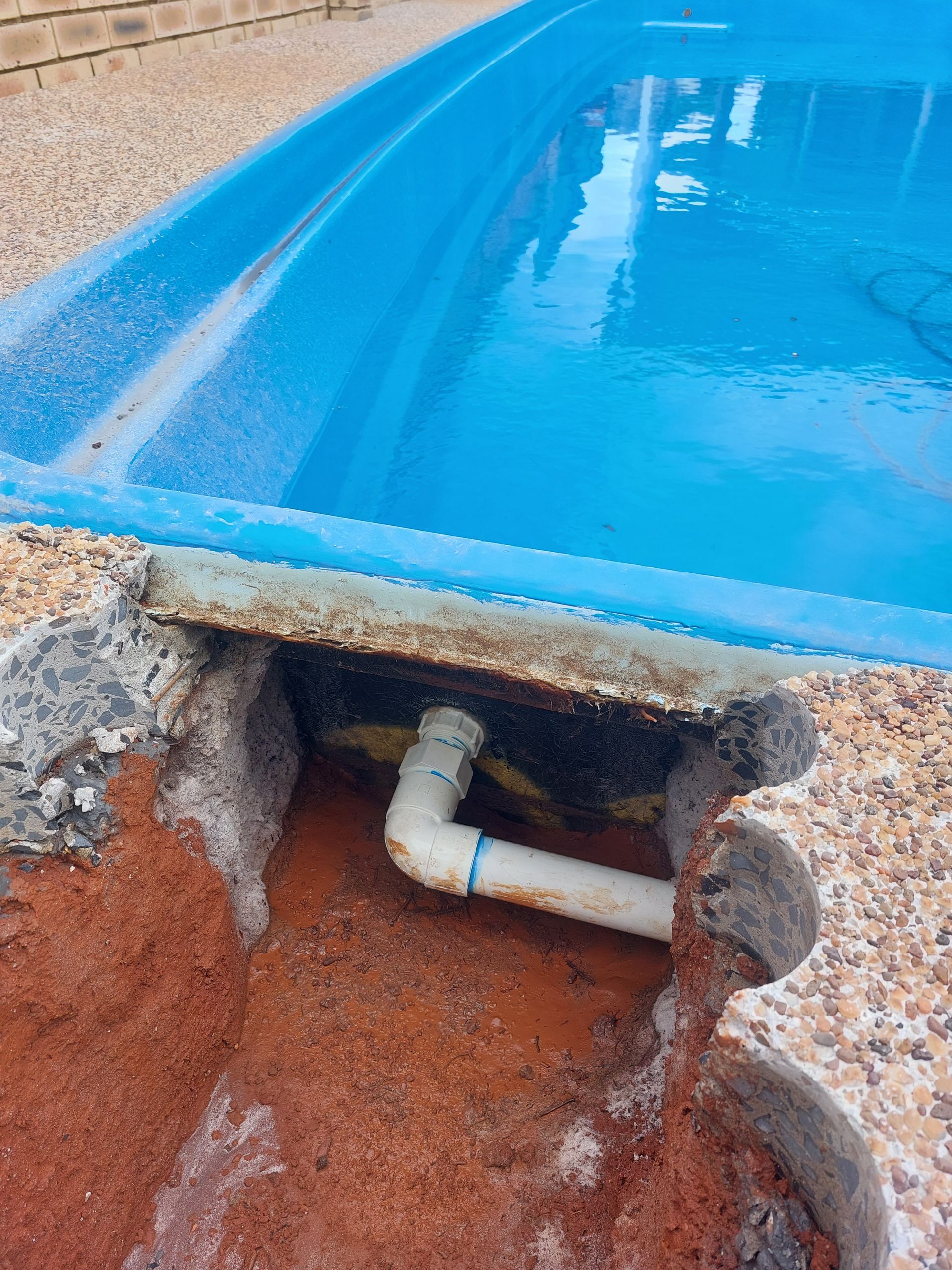 Poolside plumbing showing a white pipe connection in a weathered concrete recess; blue pool in the background.