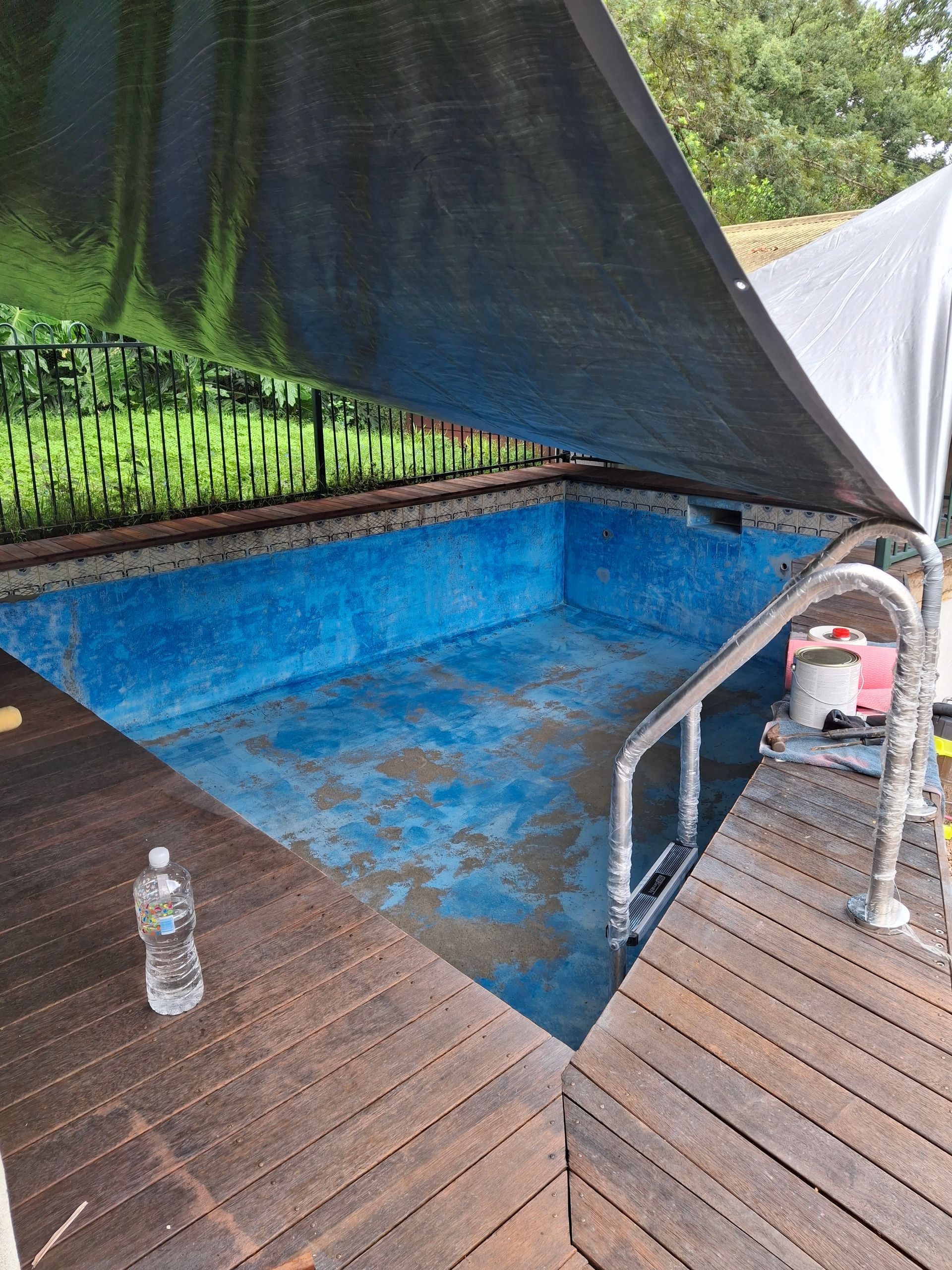 An empty, blue-lined pool with wooden decking, shaded by a dark canopy. A water bottle rests on the deck.