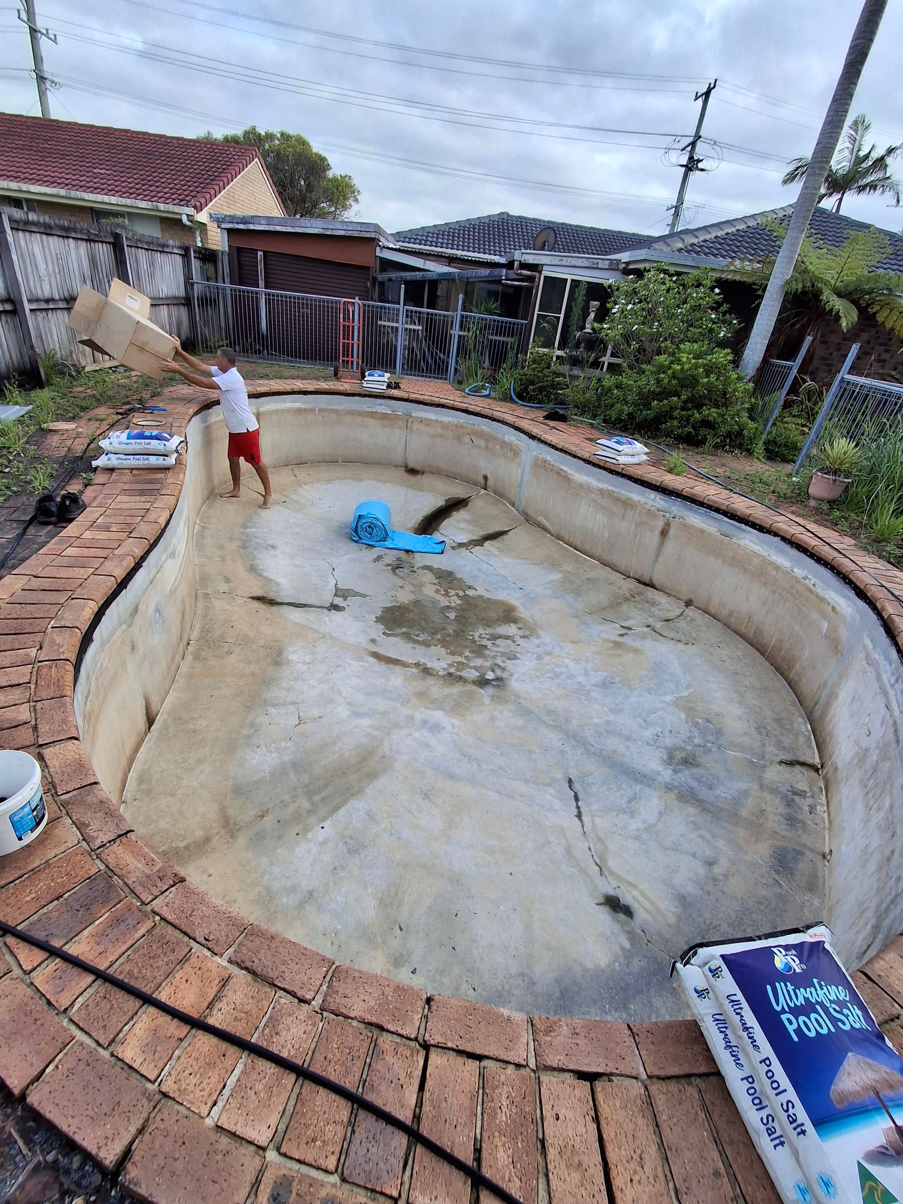 Empty, kidney-shaped pool with red brick edging. A person works inside. Blue tarp and bag of pool product visible.