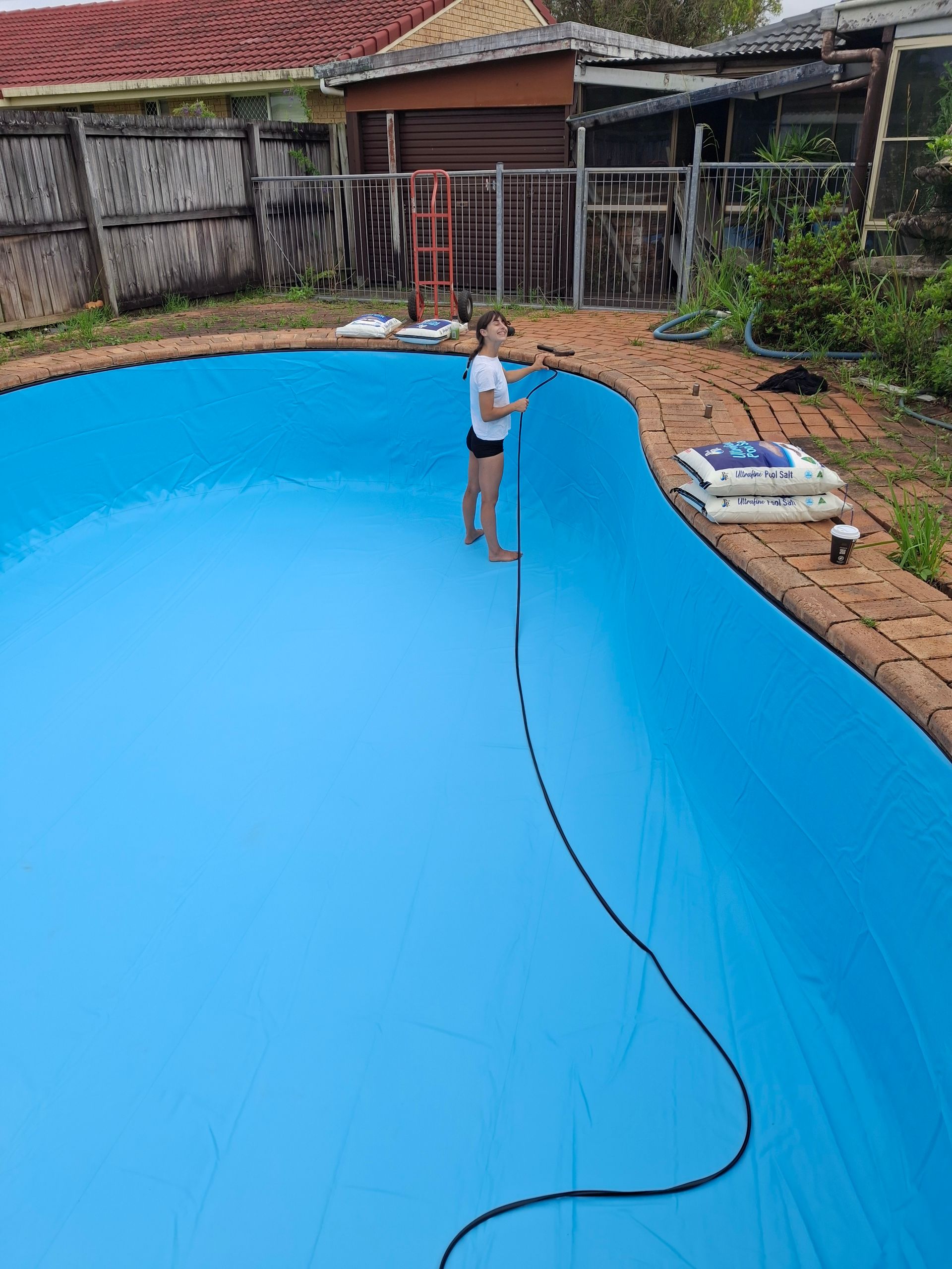 Person standing on blue pool cover, holding a hose. Bags sit nearby on brick coping. Backyard setting.