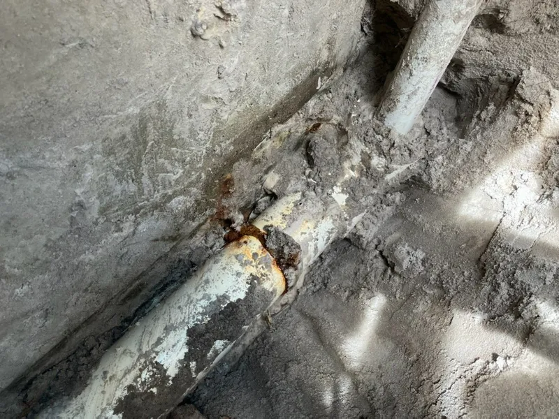 Corroded metal pipes embedded in concrete; a section is broken, exposing rust.