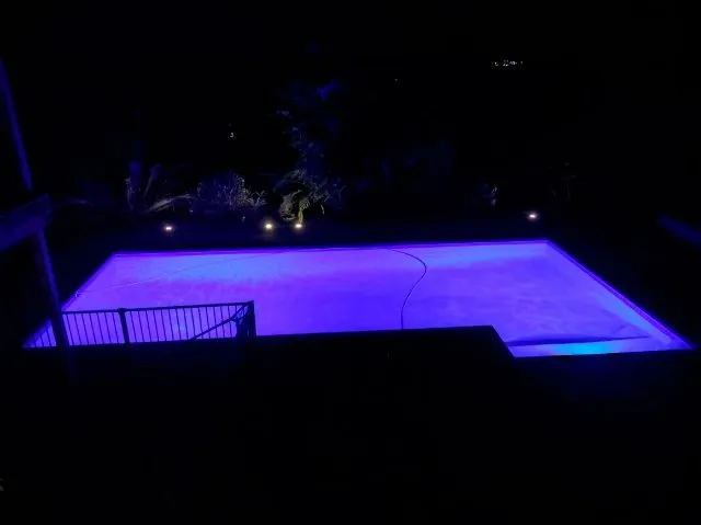 A rectangular swimming pool illuminated with purple lights at night.