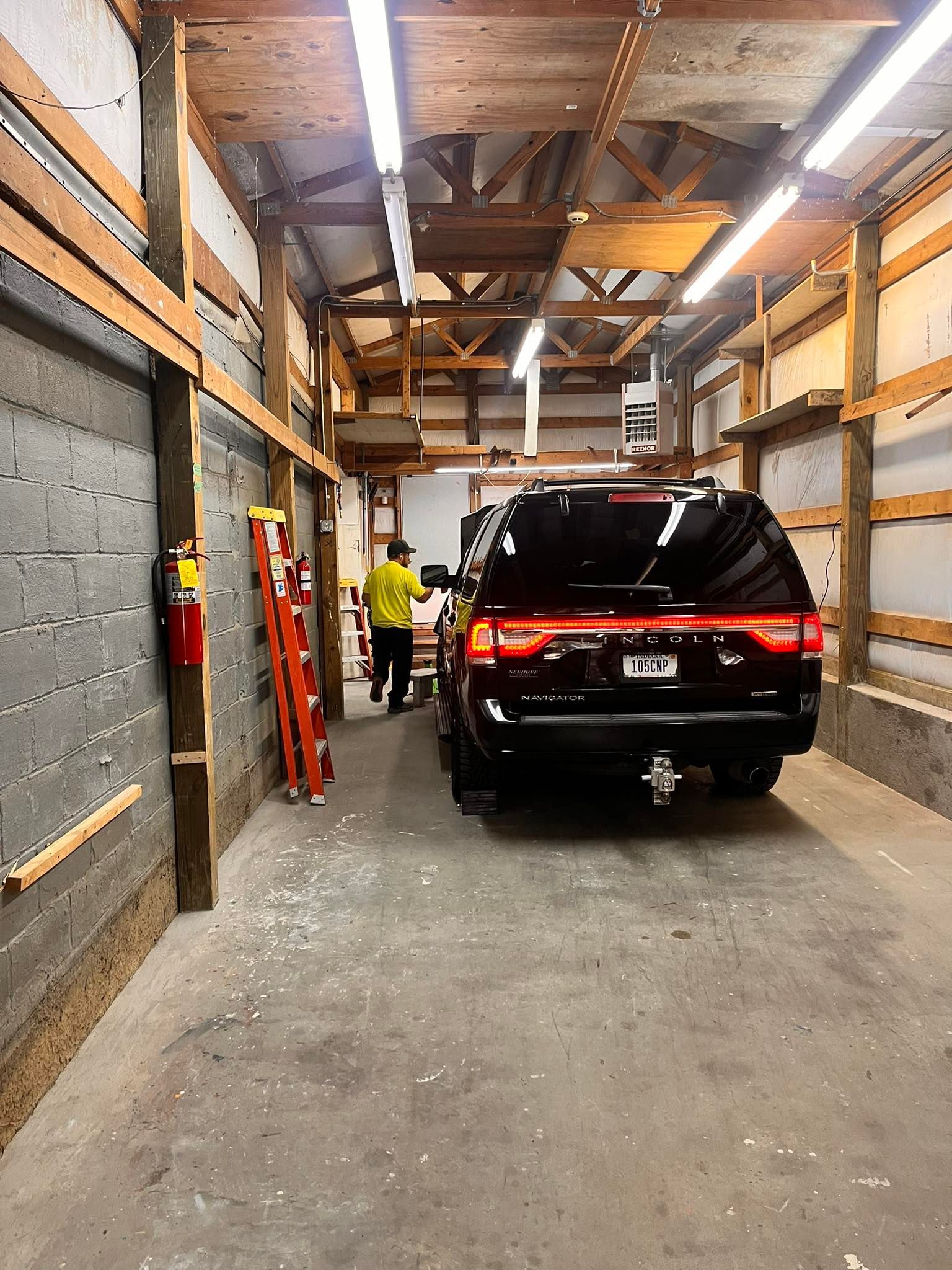 Black SUV parked in a dimly lit garage. A person walks near the vehicle. A fire extinguisher and ladder are visible.