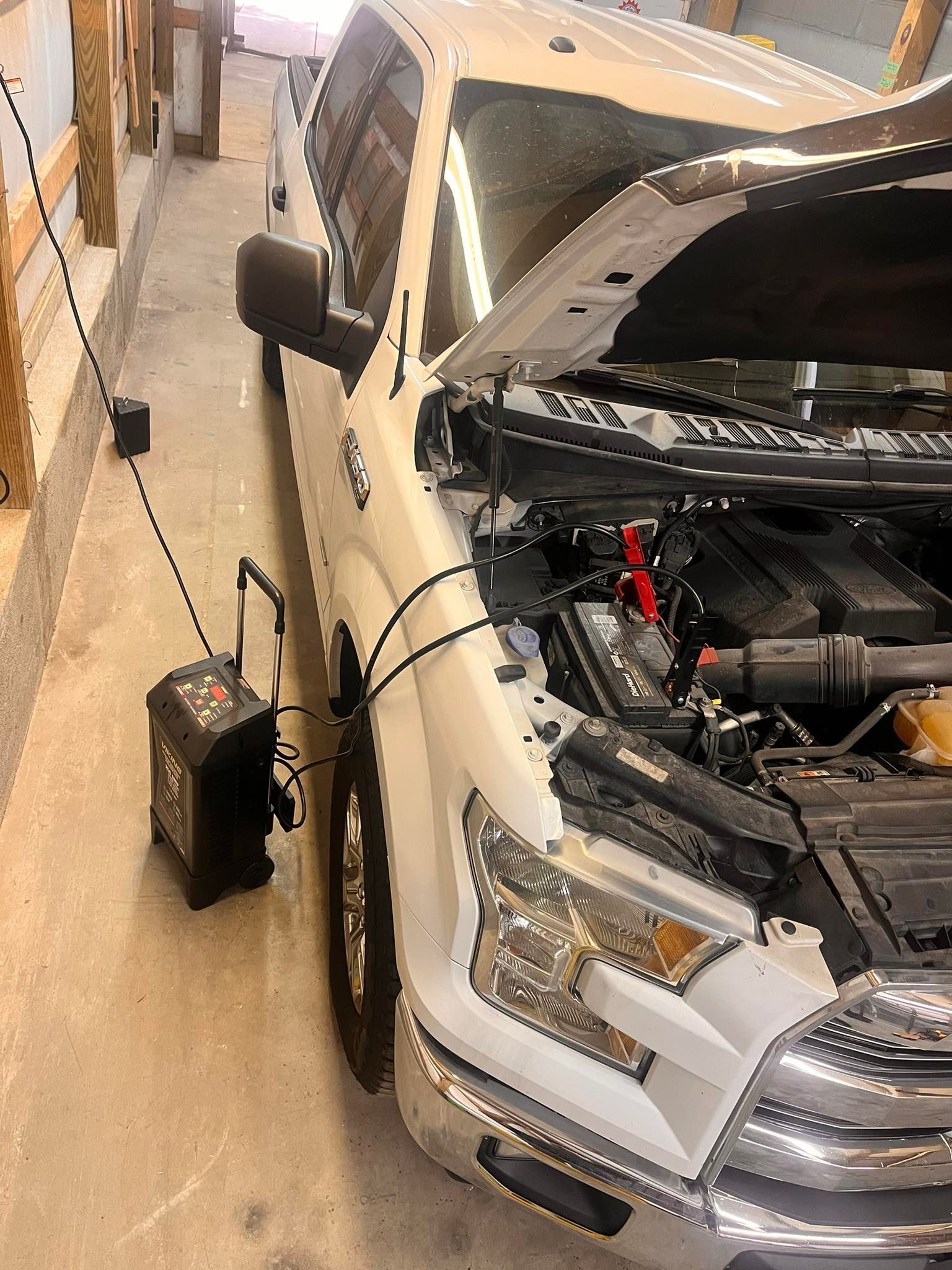 White truck with open hood, being charged by a black battery charger in a garage.