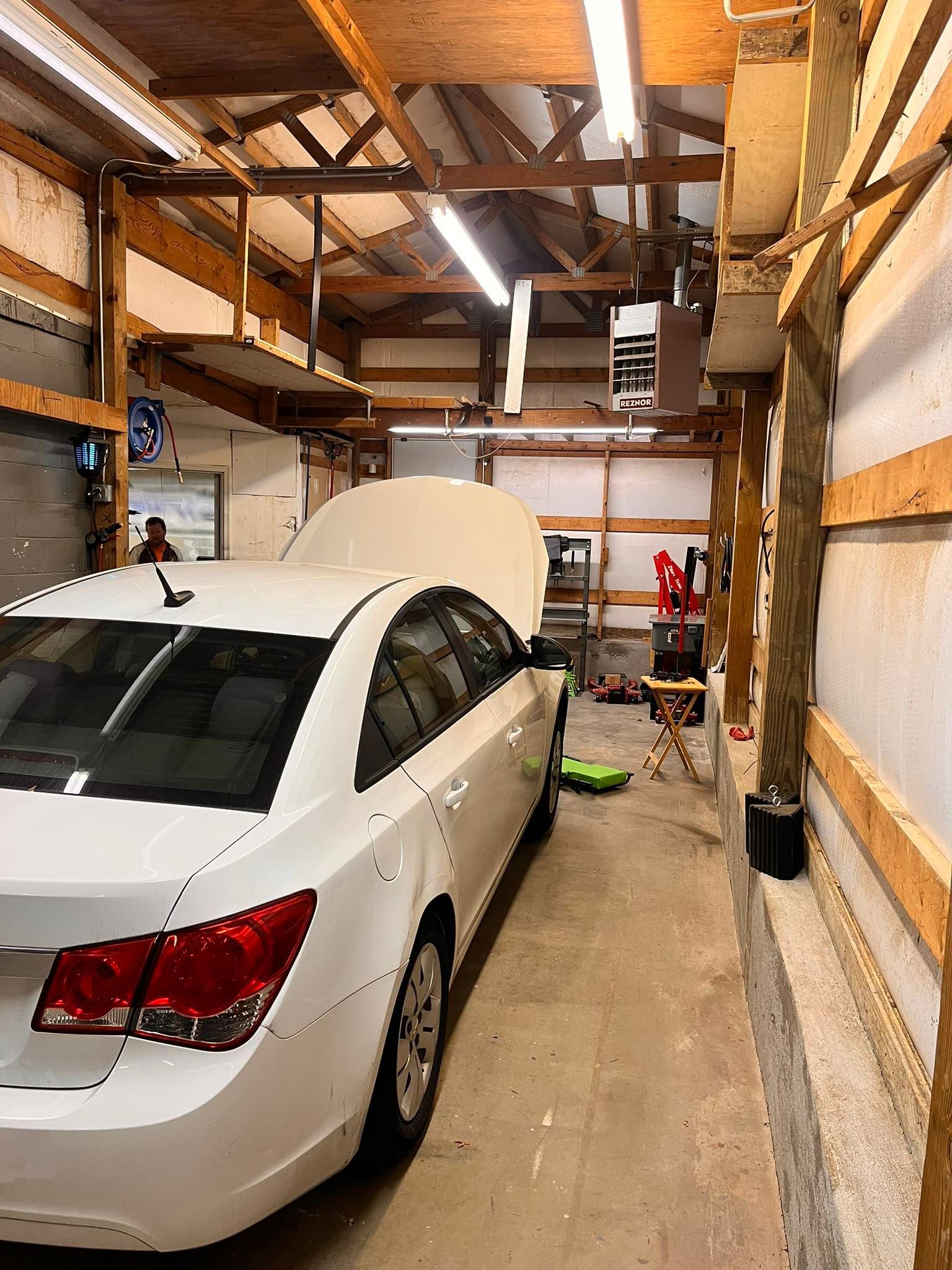 White car parked in a garage with open hood; garage interior with exposed beams and tools.