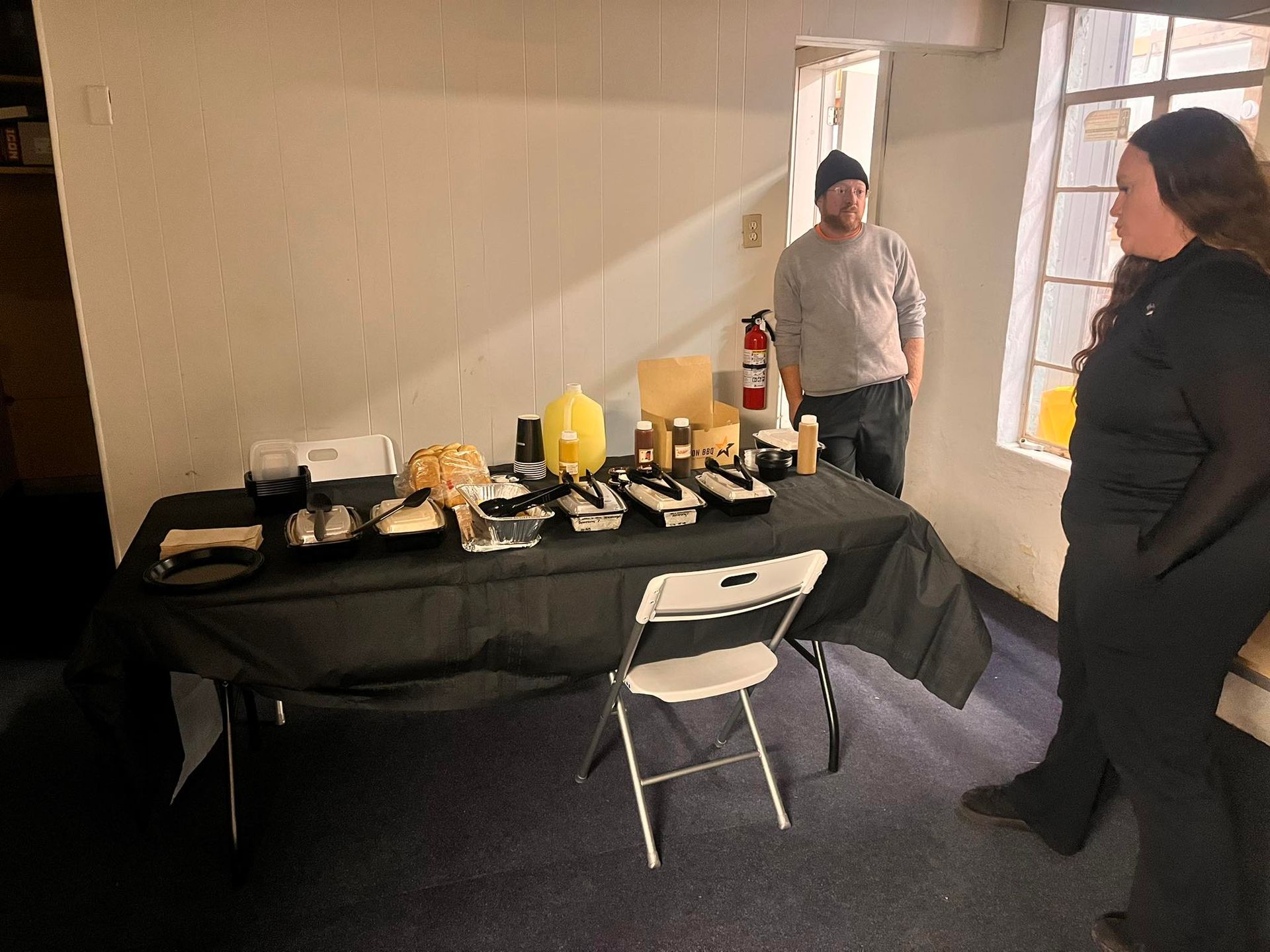 A woman and a man in a room with products on a table, including containers and a white folding chair.