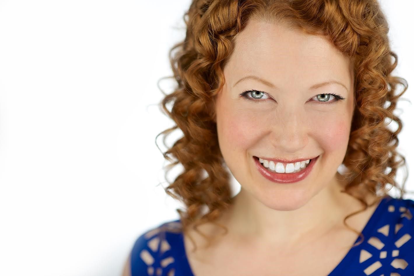 Woman with red curly hair smiling widely in a blue dress.