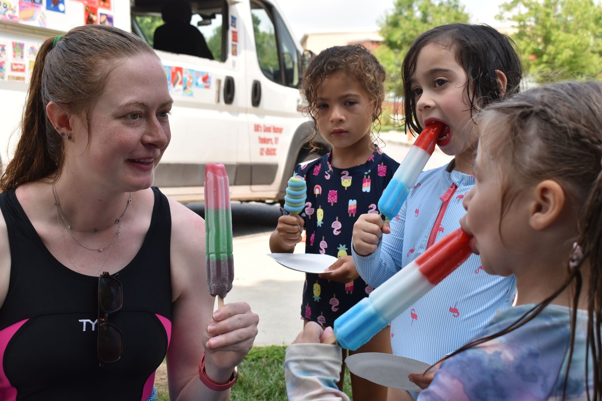 A woman in a swim suit is standing next to three little girls eating popsicles.