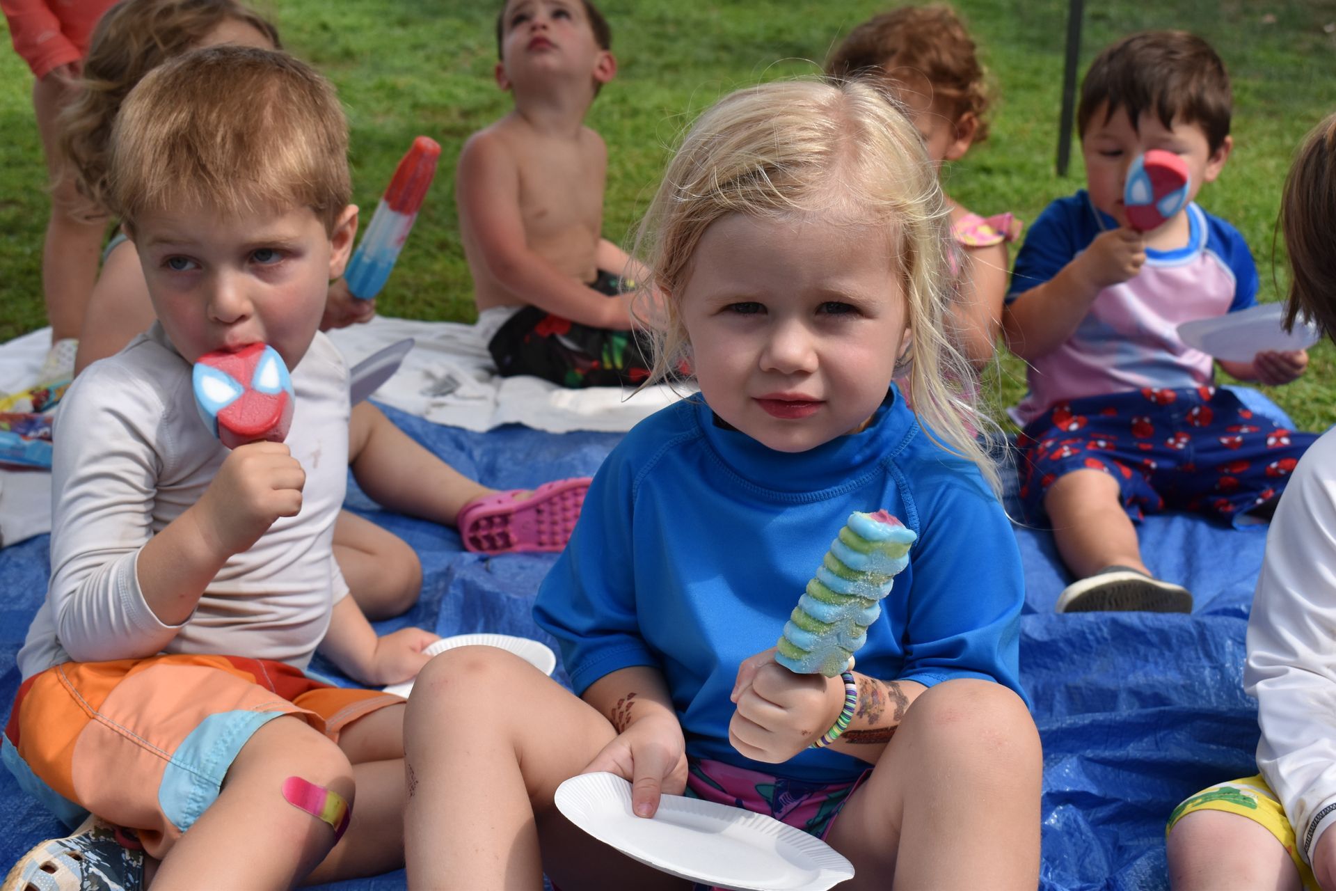 A group of children are sitting on a blue tarp eating popsicles.