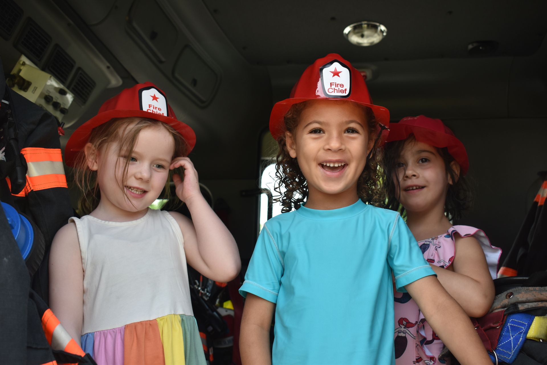 Three little girls wearing firefighter hats are standing in a fire truck.