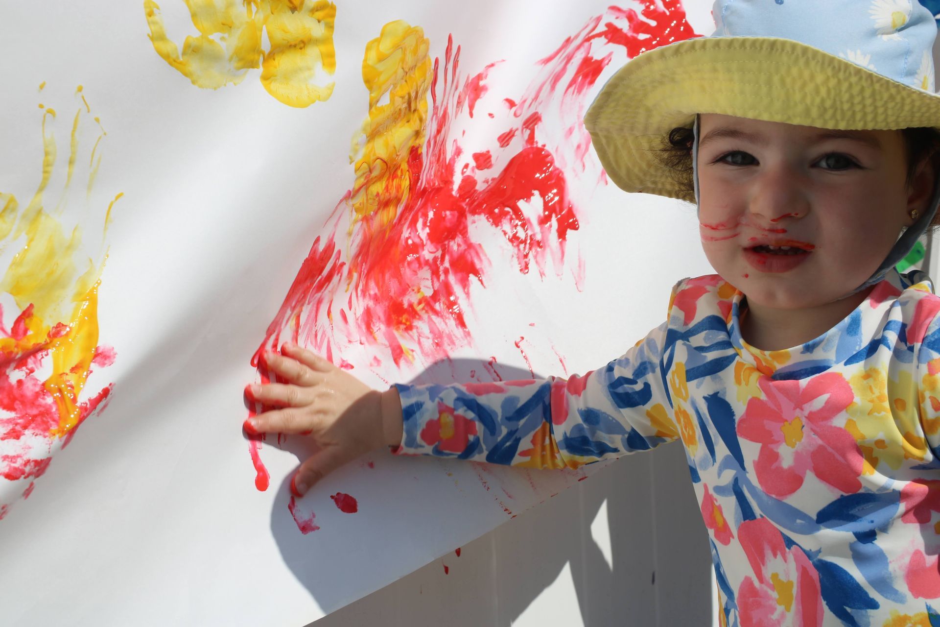 A little girl wearing a hat is standing in front of a wall covered in paint.