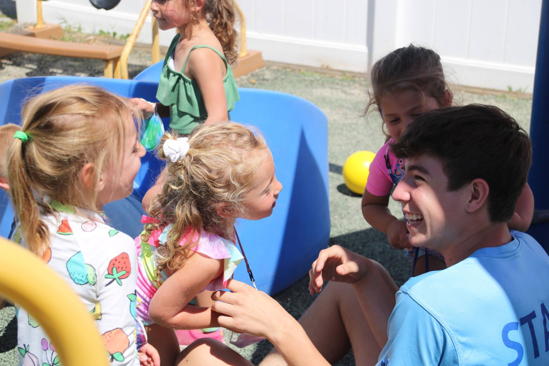 A man wearing a blue shirt that says ' st ' on it is talking to a group of children