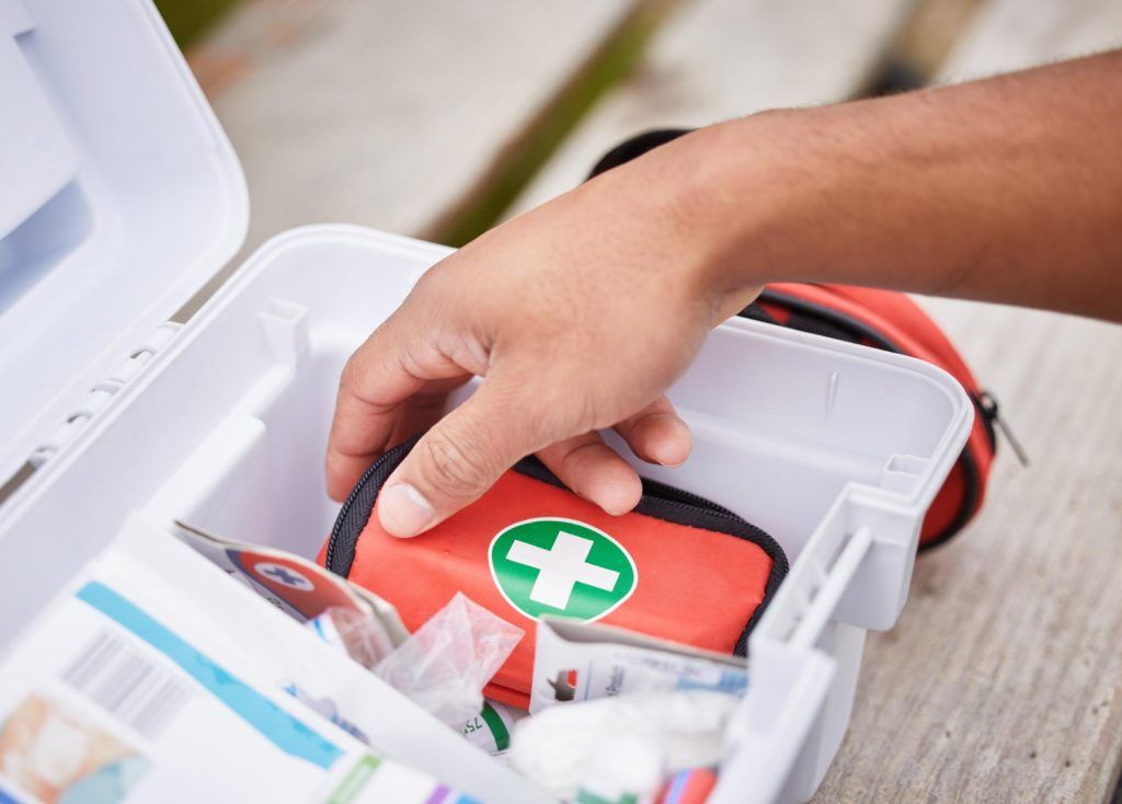 Hand reaching into a white first-aid kit to retrieve a small red bag with a green cross symbol.
