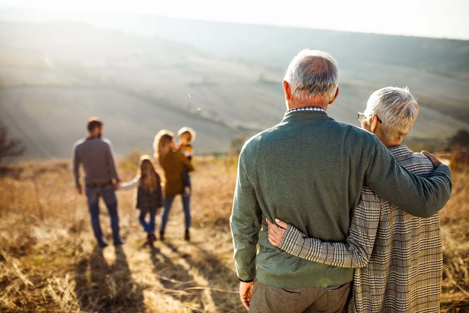 Older couple embraces, looking at a family in a sunlit field; countryside background.