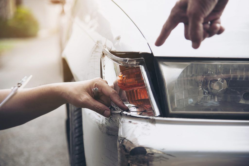 Hands pointing to damage on a car's headlight and bumper, likely assessing the accident's aftermath.