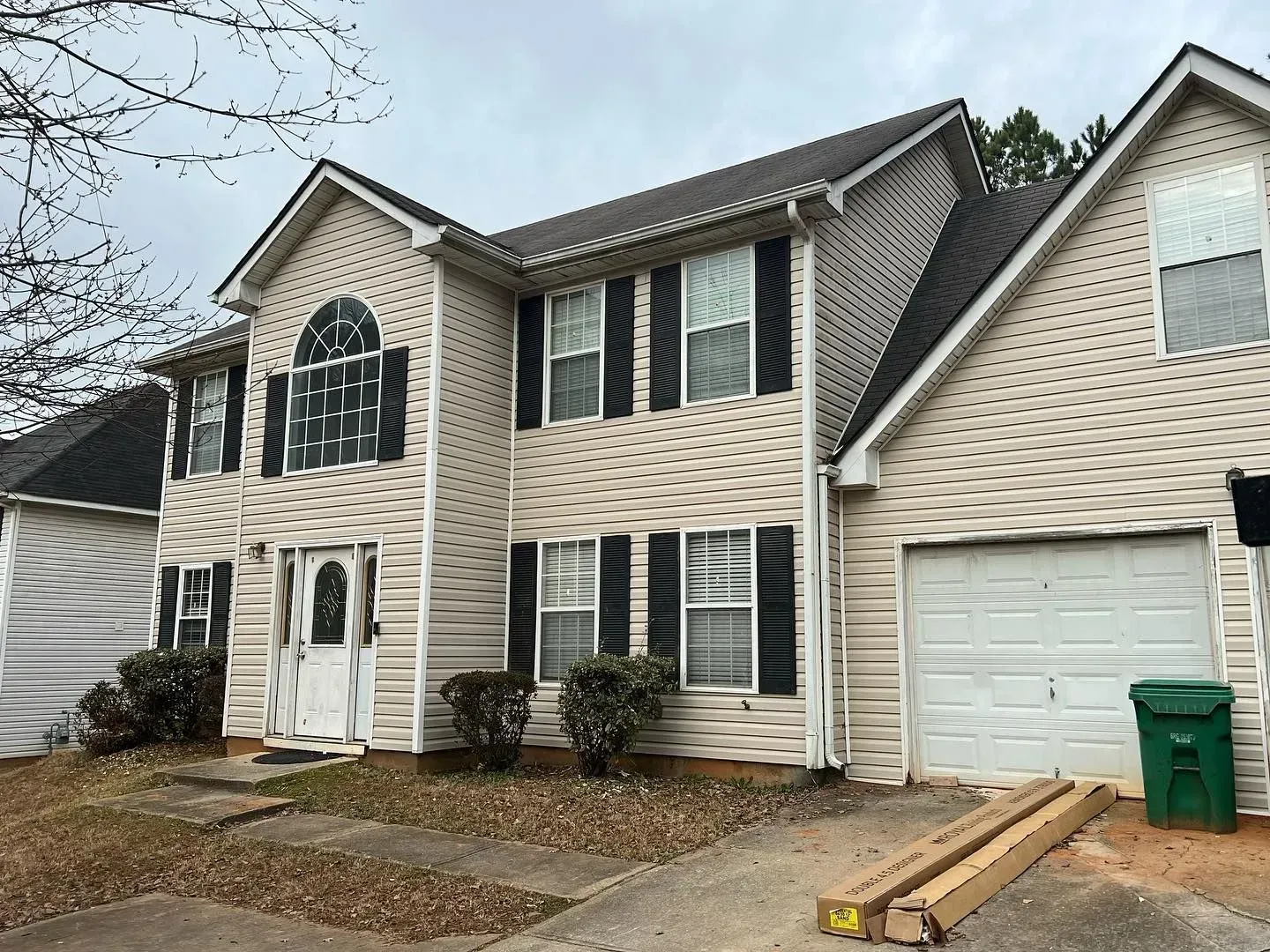 A large white house with black shutters and a green trash can in front of it.