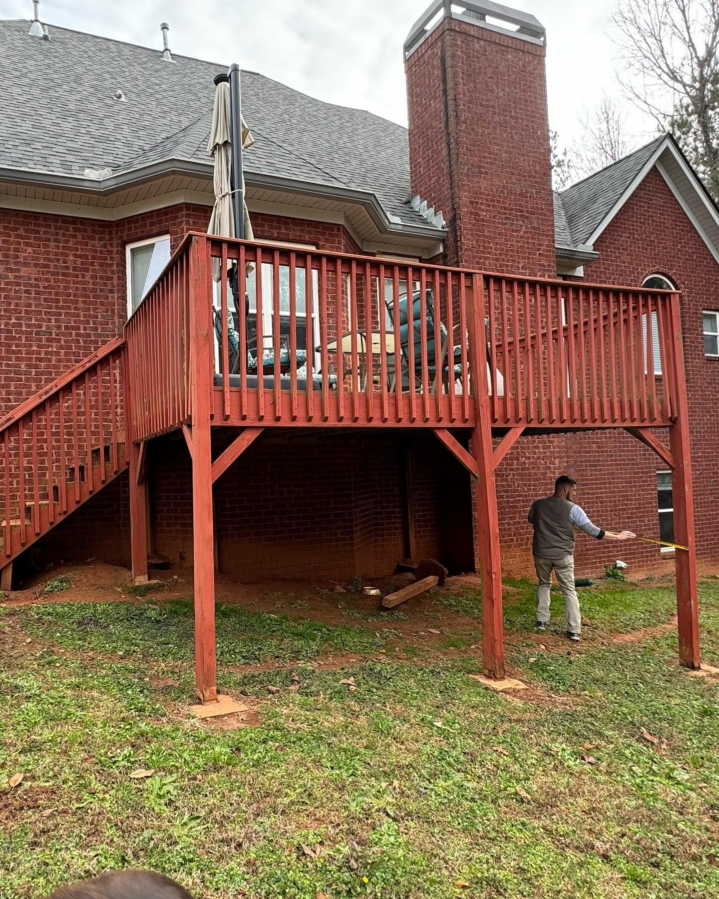 A man is standing next to a wooden deck in front of a brick house.