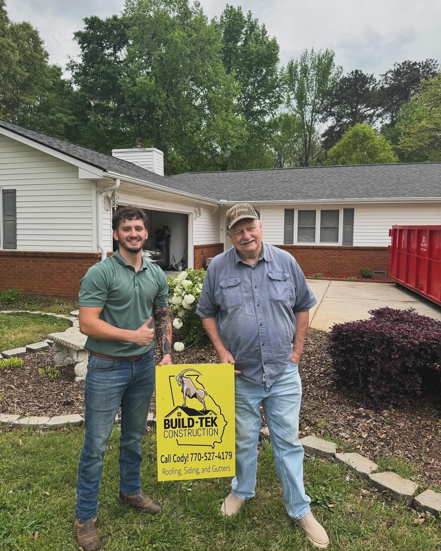 Two men are standing in front of a house holding a sign.