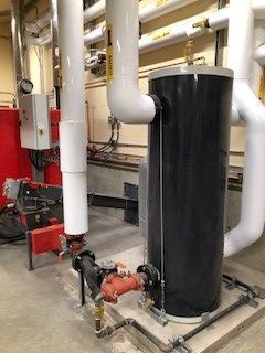 A black water heater is sitting in a room surrounded by pipes.