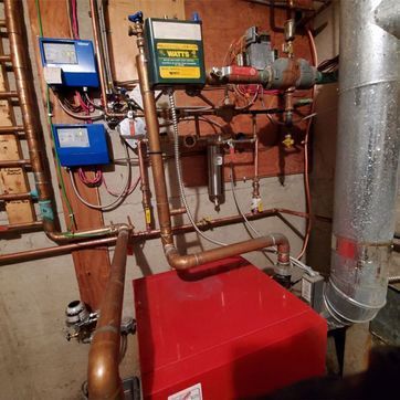 A red boiler is sitting in a basement surrounded by pipes.