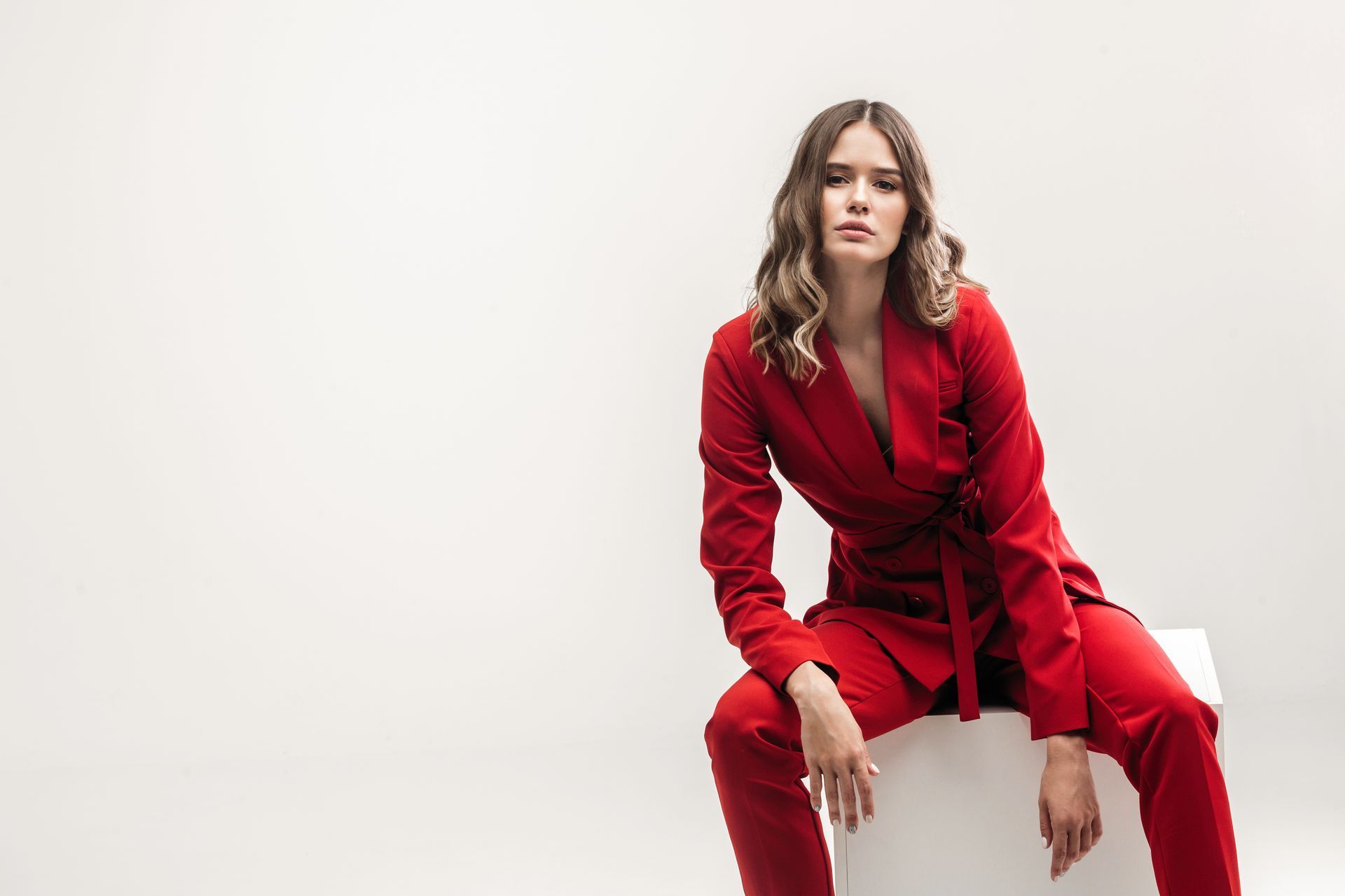 A woman in a red suit is sitting on a white cube.