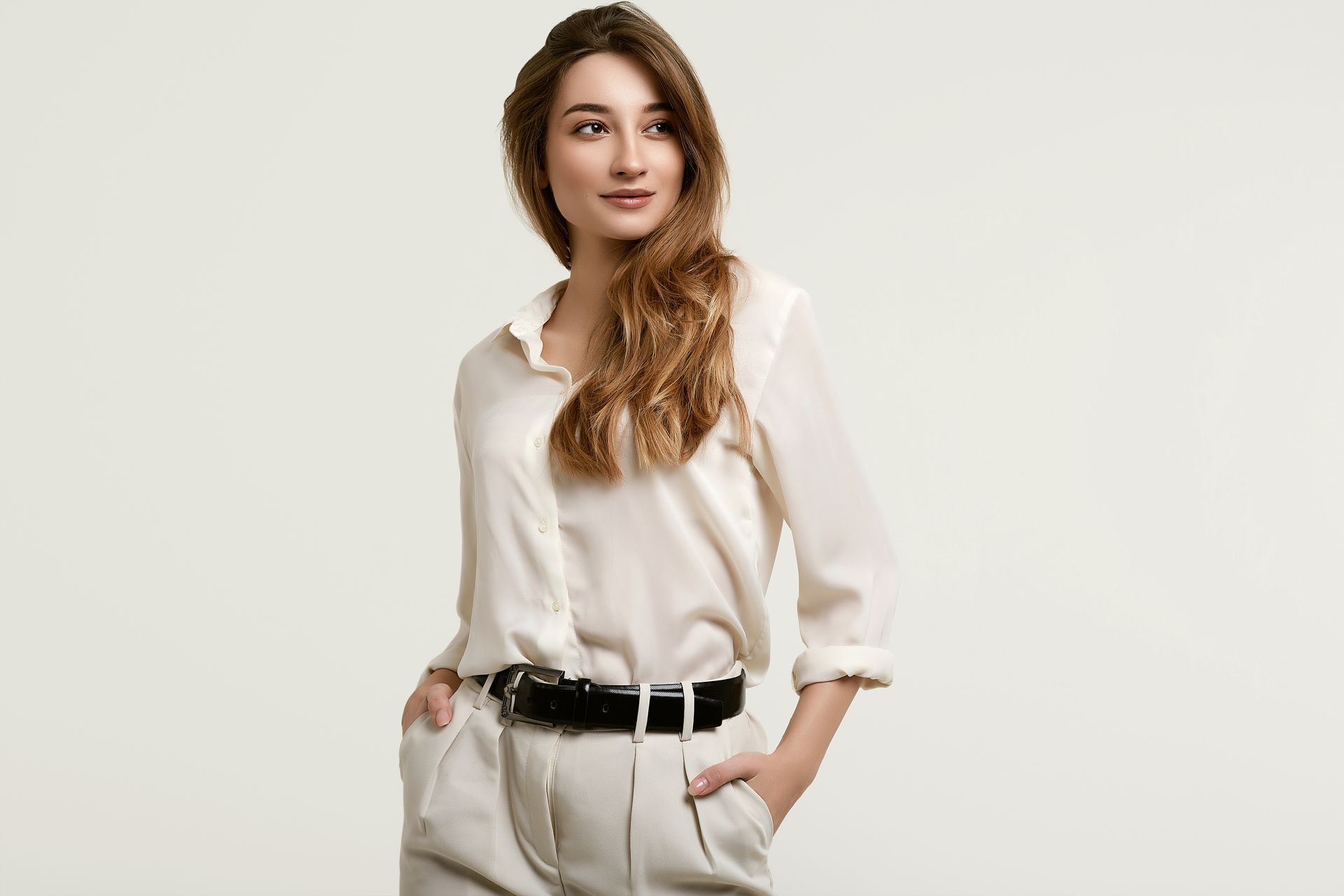 A woman in a white shirt and beige pants is standing with her hands in her pockets.