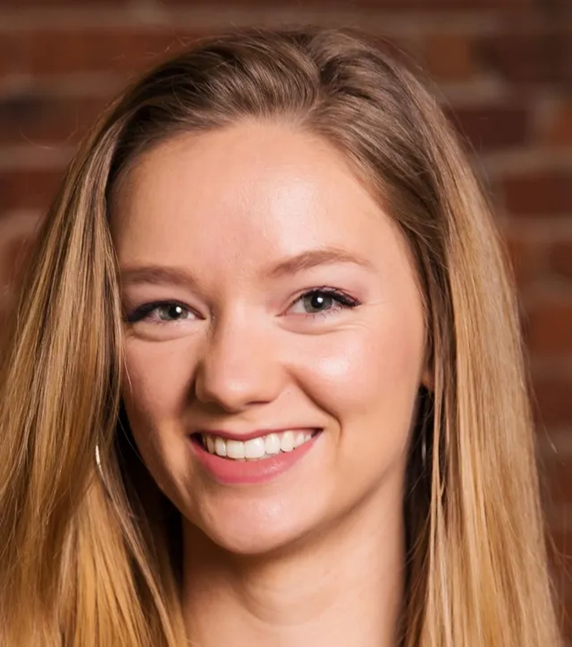 A woman with long blonde hair is smiling in front of a brick wall.