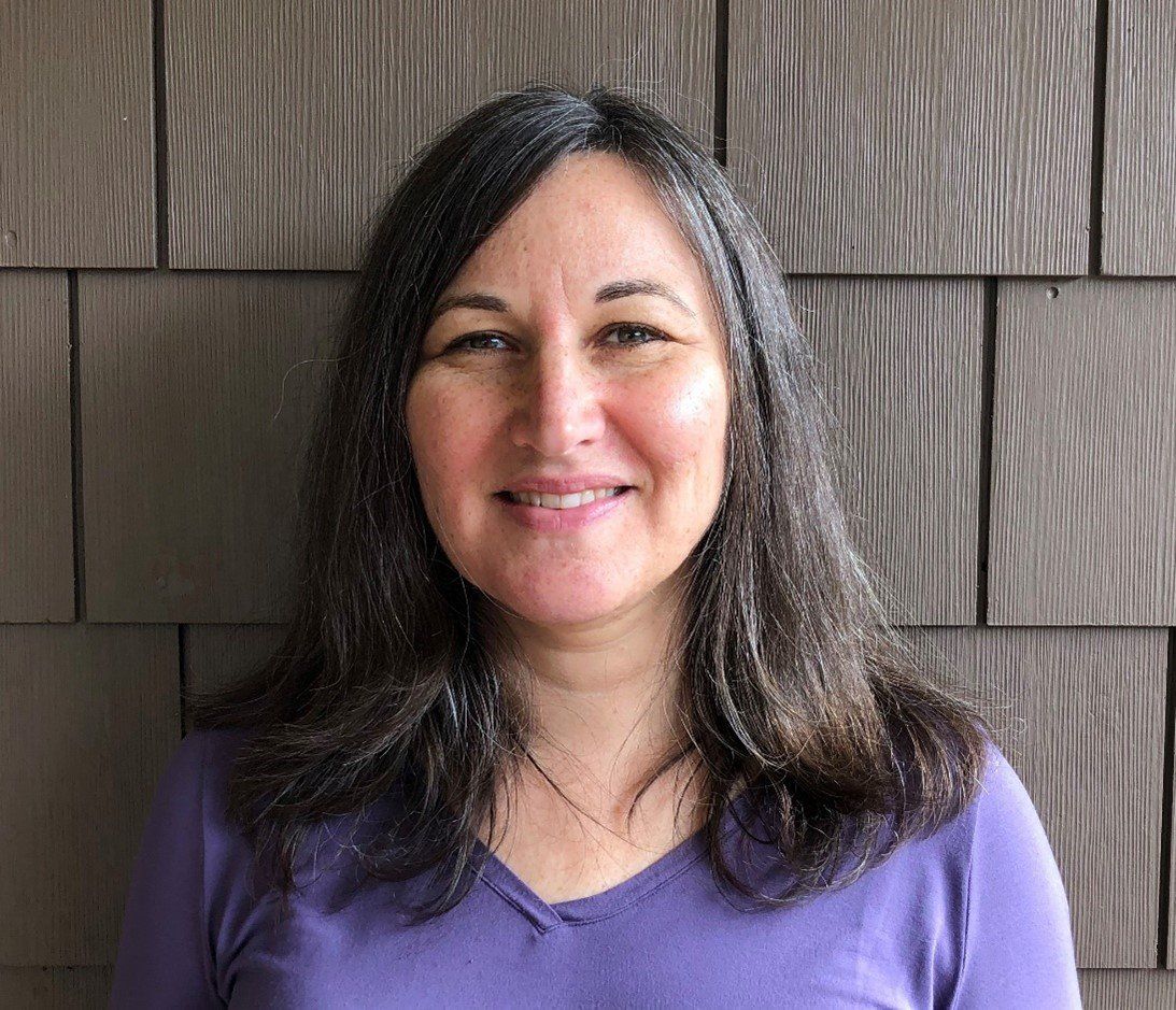 A woman in a purple shirt is smiling in front of a wooden wall.