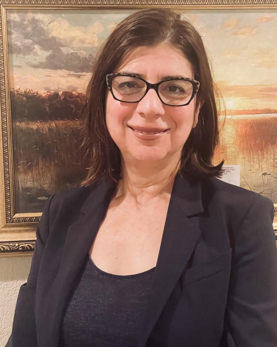 A woman wearing glasses and a black jacket is smiling in front of a painting.