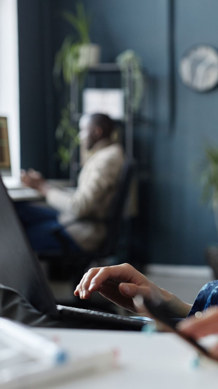 Person typing on laptop in focus; another person blurred in background working at a desk. Dark room, plants.