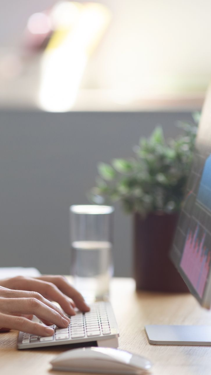 Hands typing on a keyboard, with a computer monitor, plant, and glass of water on a desk.