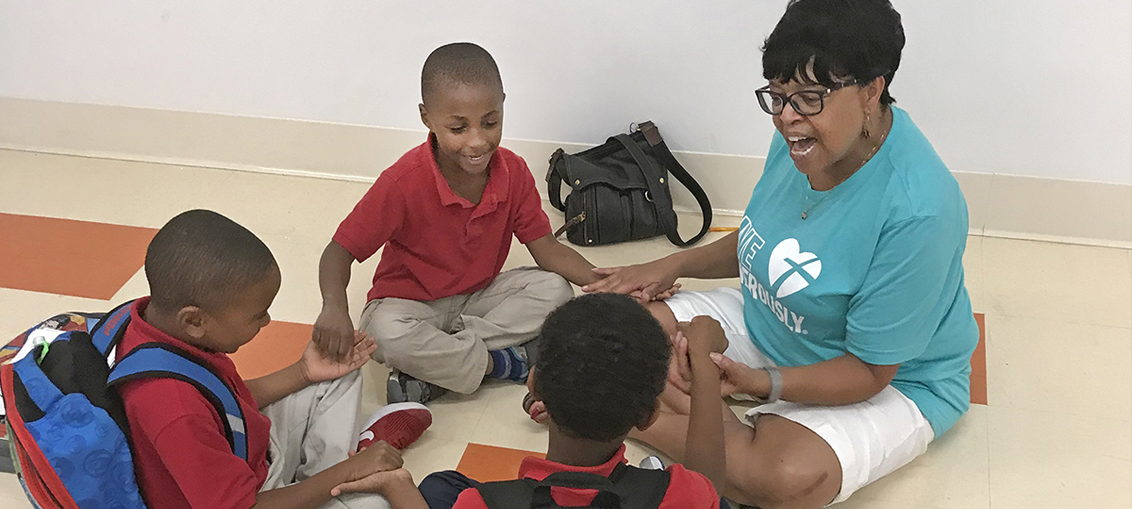 A teacher and three students sit in a circle, holding hands. The teacher wears glasses and a blue shirt.