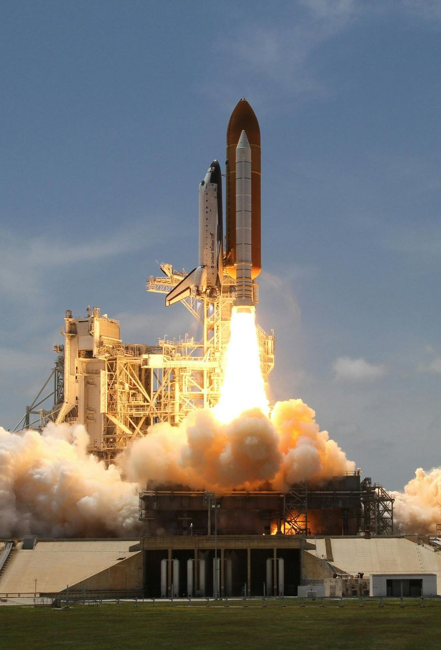 Space Shuttle launching, engulfed in fire and smoke, ascending from launch pad against blue sky.