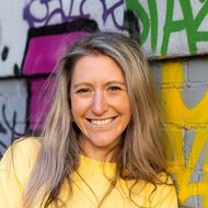 A woman in a yellow shirt is smiling in front of a graffiti wall.