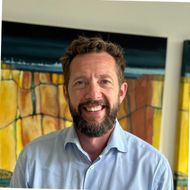 A man with a beard is smiling in front of a painting.