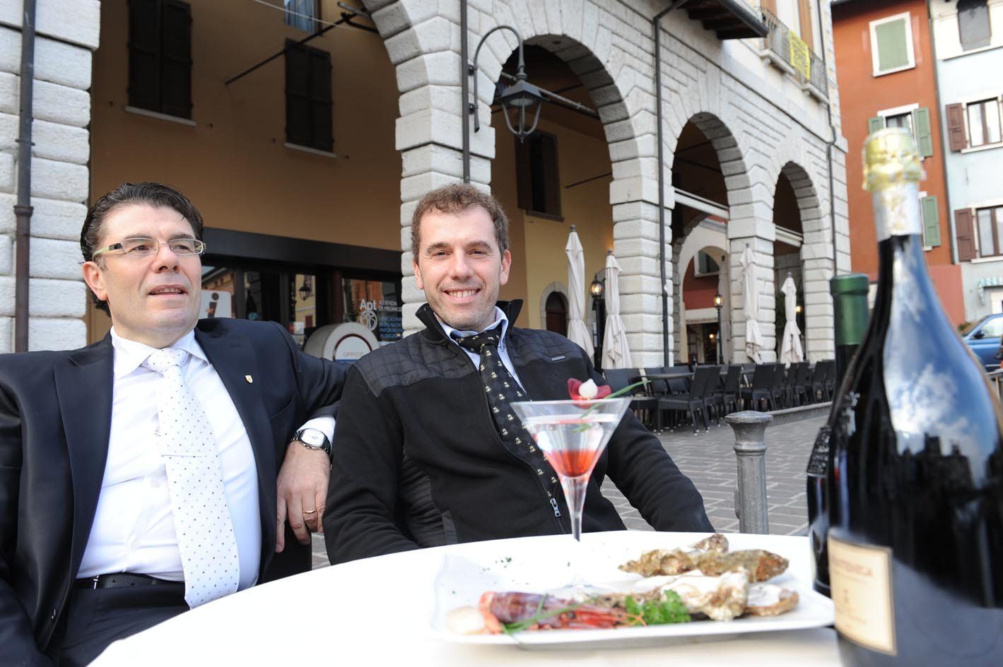 2 uomini seduti dietro un tavolo circolare bianco con cibo, bicchiere e bottiglia