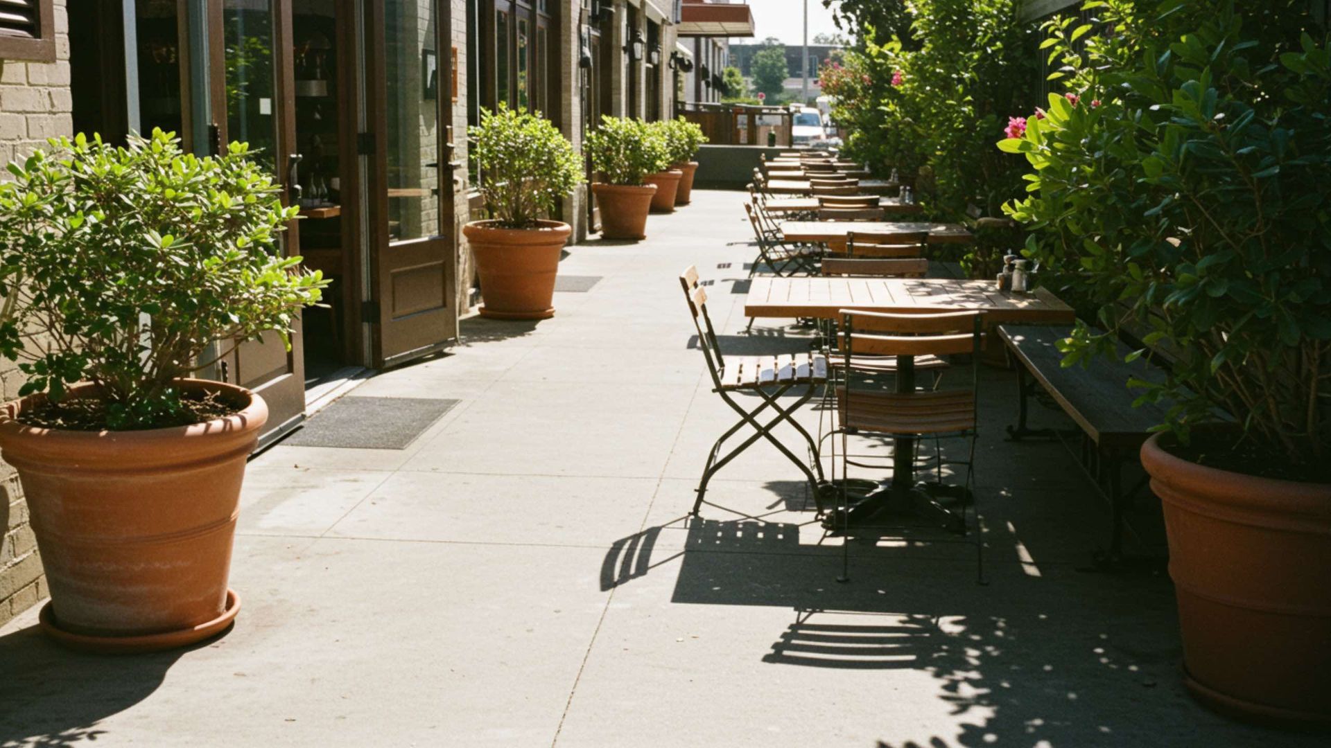 Outdoor sidewalk dining area with tables, chairs, and potted plants lined up along a brick building.