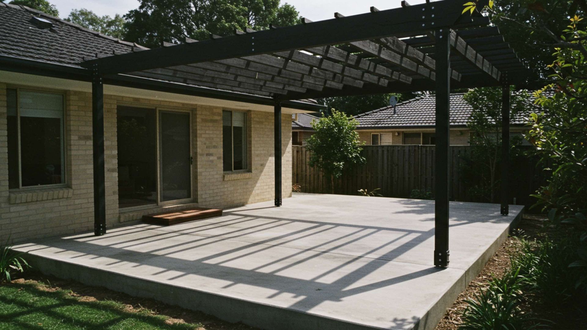 Concrete patio with a black pergola, attached to a brick house. Sunny with shadows.