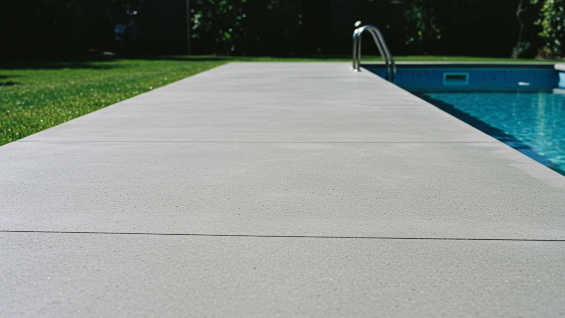 Gray concrete pool deck next to a pool with blue water and grass.