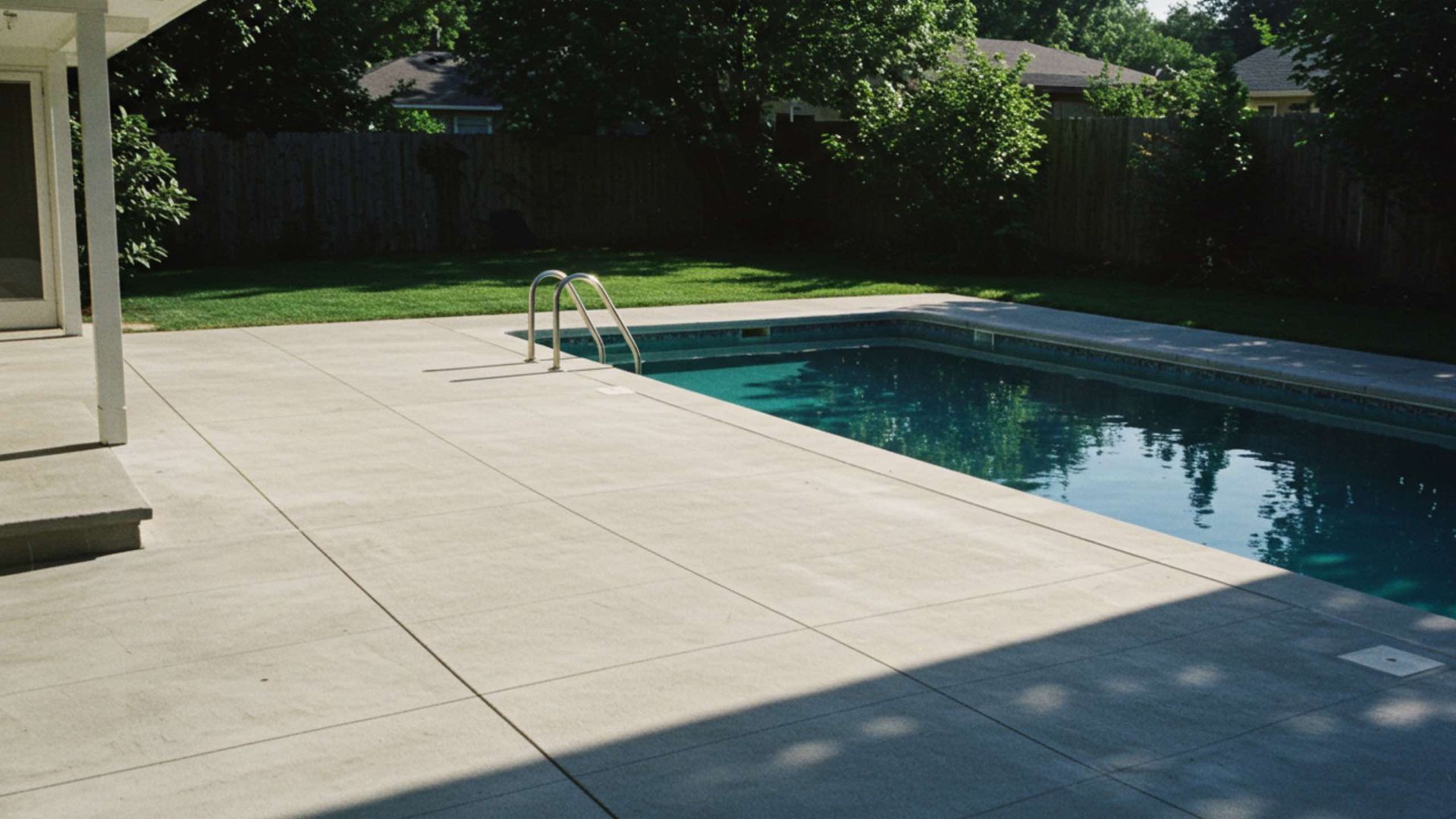 Backyard with a swimming pool and concrete patio. Green grass, trees, and a wooden fence are in the background.