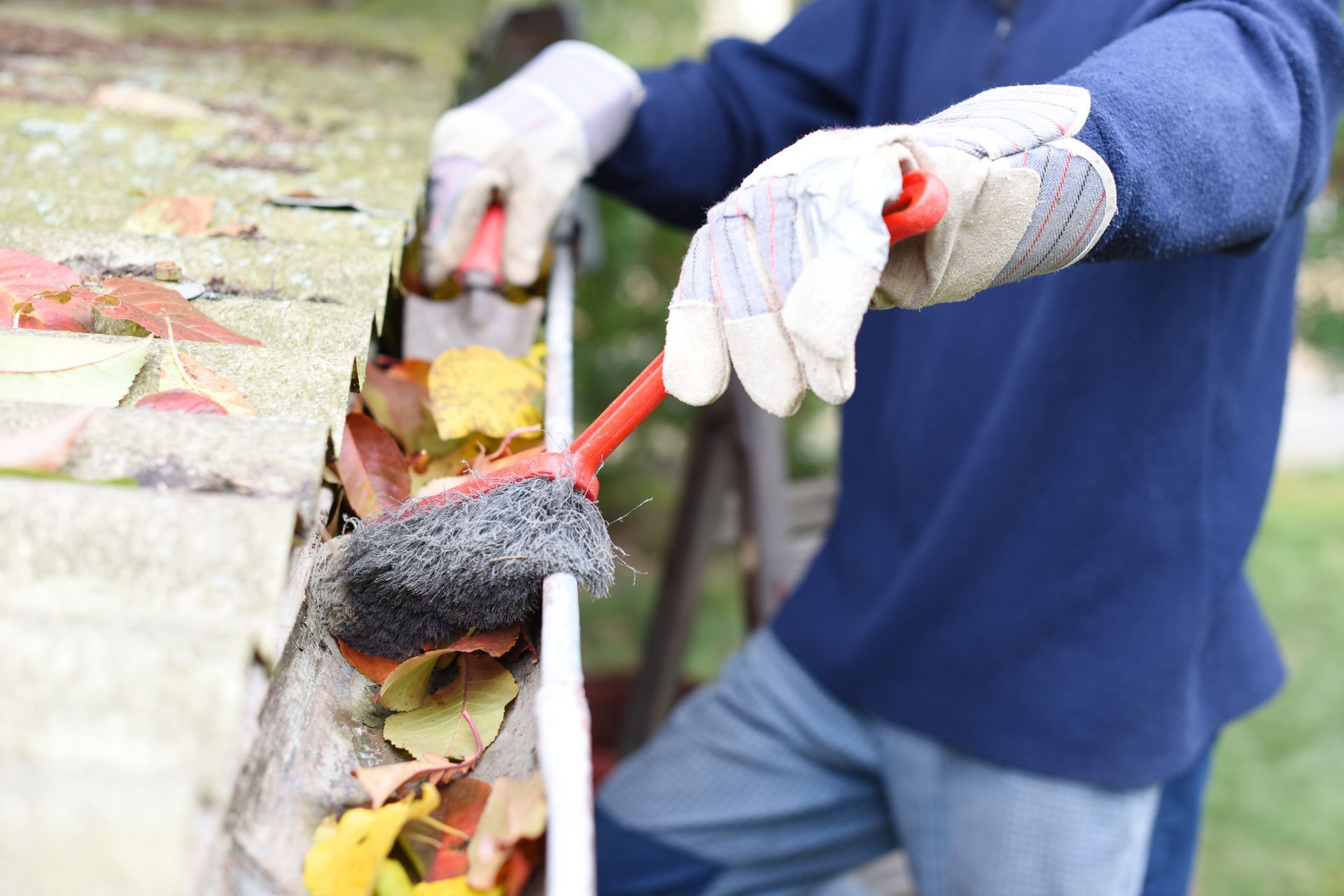 Person clearing leaves from a gutter with a rake and gloved hands.