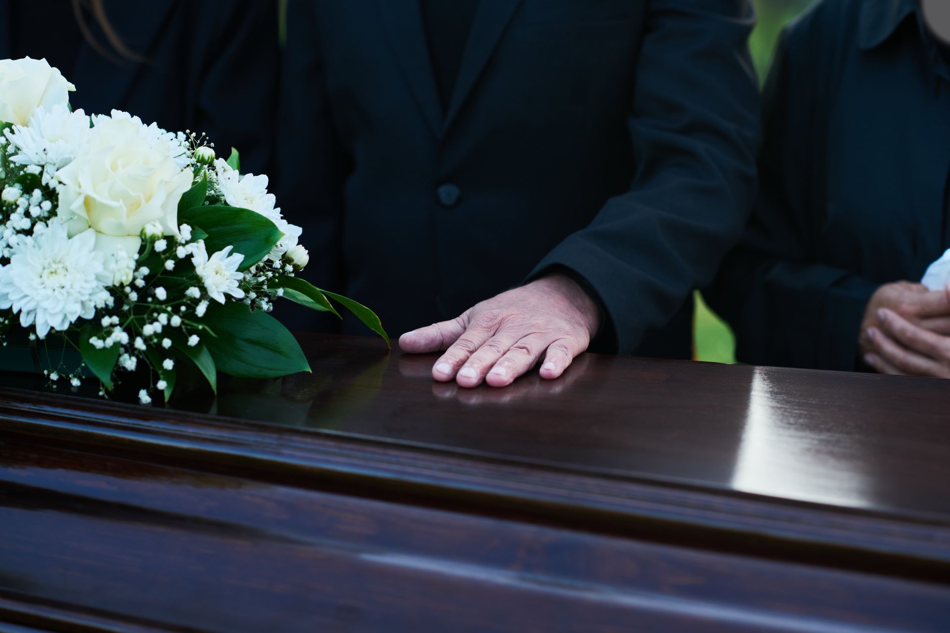 Hands resting on a polished casket beside white flowers at a funeral