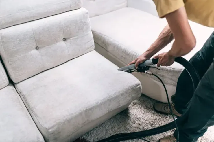Upholstery cleaner vacuuming a light gray sofa and ottoman in a living room