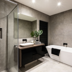 Modern bathroom with gray stone walls, glass shower, floating vanity, and white bathtub with black towel