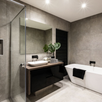 Modern bathroom with gray stone walls, glass shower, floating vanity, and white bathtub with black towel