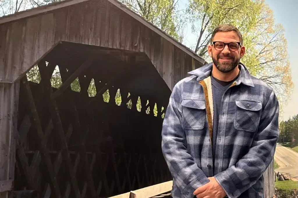 A man in a plaid shirt is standing in front of a covered bridge.