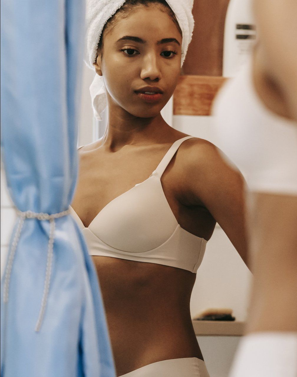 A woman in a bra with a towel wrapped around her head is looking at herself in the mirror.