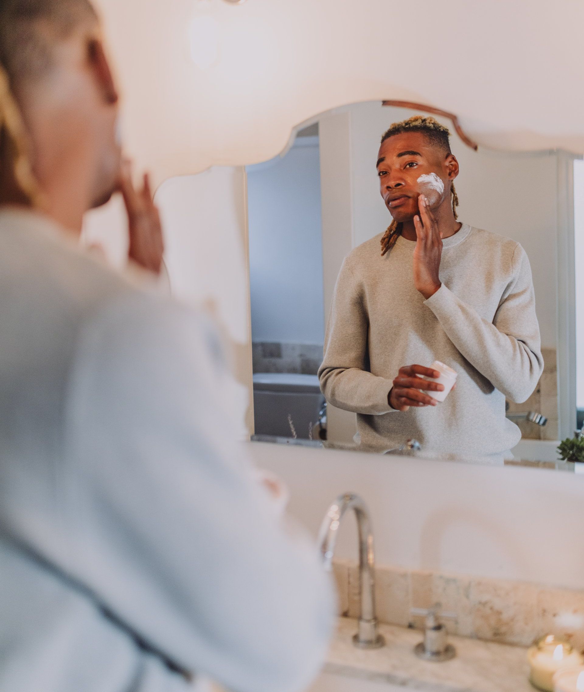 A man is applying cream to his face in front of a bathroom mirror.