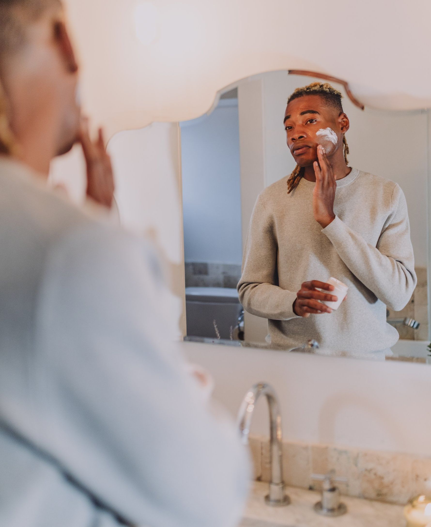 A man is applying cream to his face in front of a bathroom mirror.