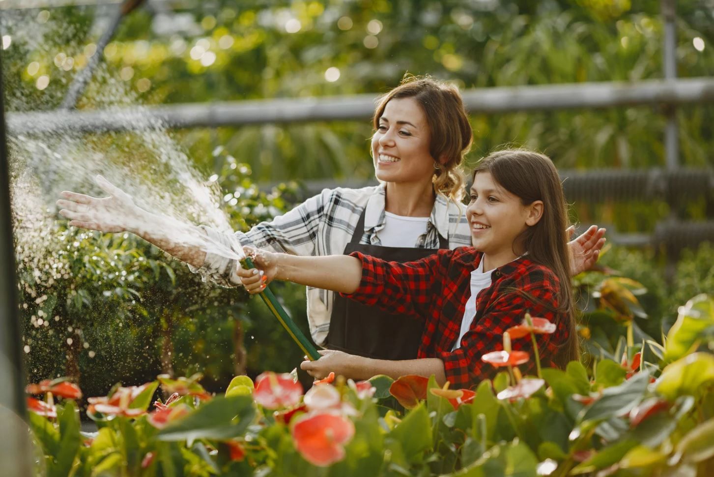 A woman and a girl are watering flowers with a hose.